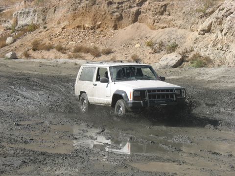 1987 Ford Bronco - A white Jeep Cherokee XJ navigates a muddy off-road trail, kicking up water and dirt.
