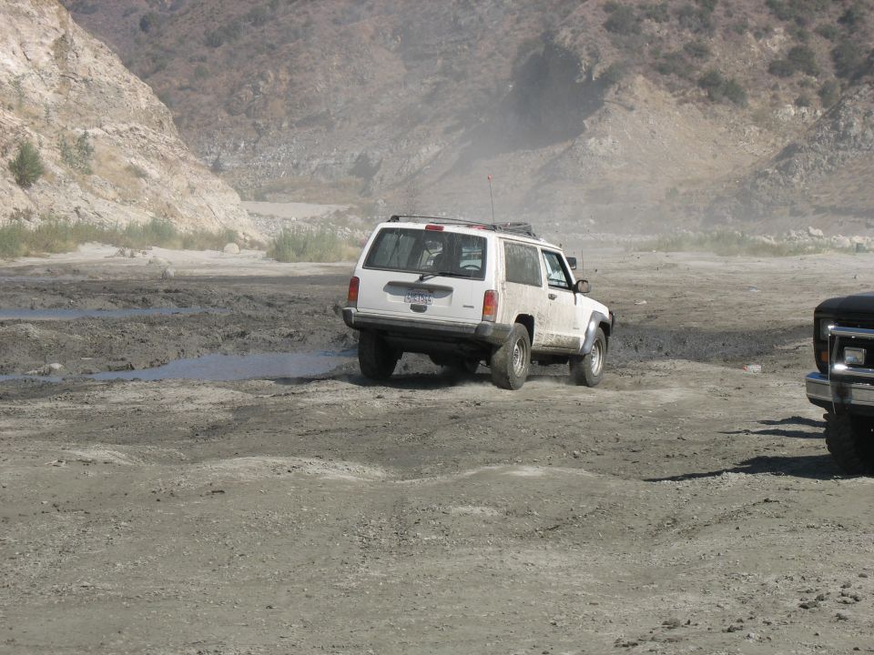 White SUV driving on a dusty, muddy dirt road with mountains in the background. Another vehicle is partially visible on the right