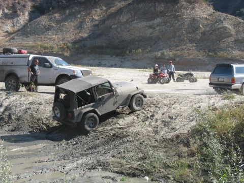 1987 Ford Bronco - Multiple off-road vehicles navigate a muddy terrain, with a Jeep Wrangler appearing to be stuck.