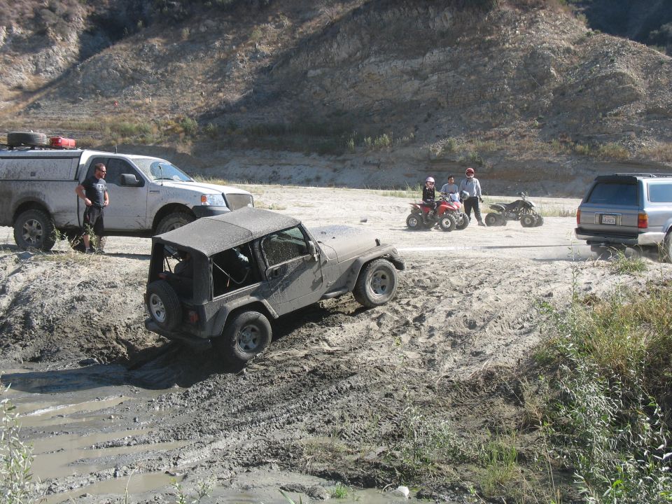 Jeep Wrangler stuck in mud with a Ford F-150, Toyota 4Runner, and ATVs nearby