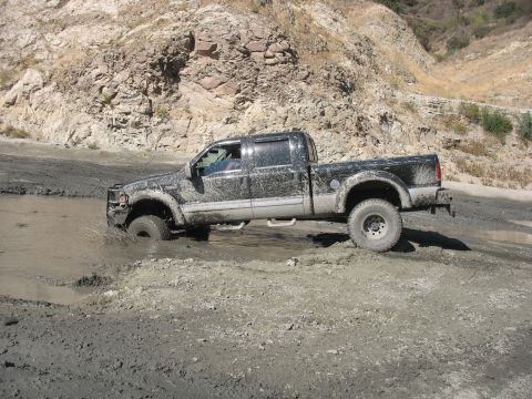 1987 Ford Bronco - A four-wheel drive pickup truck navigates through a muddy terrain, with a rocky hillside providing a backdrop.