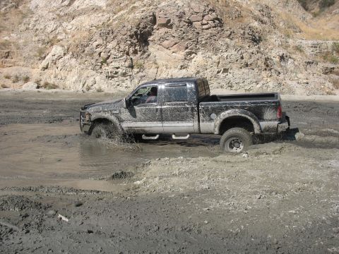 1987 Ford Bronco - A black pickup truck navigates through a muddy terrain, with water and mud spraying around its wheels.
