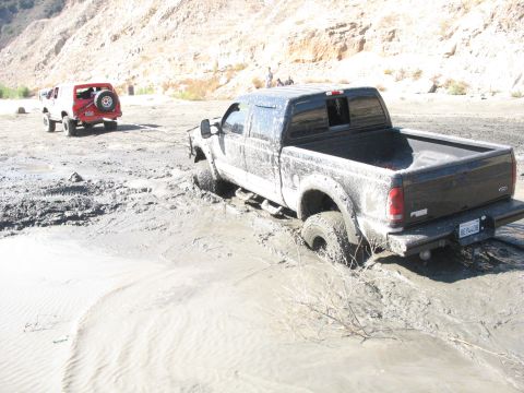 1987 Ford Bronco - A black pickup truck is stuck in thick mud, with a red SUV visible in the background.