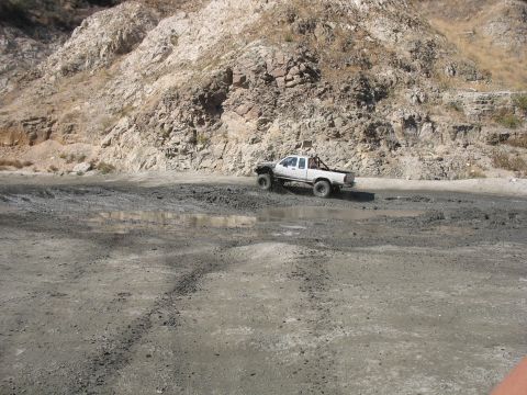 1987 Ford Bronco - Pickup truck navigates a muddy track with a rocky mountain backdrop.