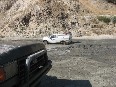 1987 Ford Bronco - A white pickup truck with a utility cap is stuck in thick mud on a dirt road, with another vehicle partially visible in the foreground.