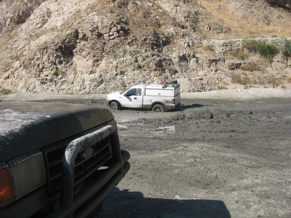 White pickup truck with a utility cap stuck in mud, viewed from another vehicle