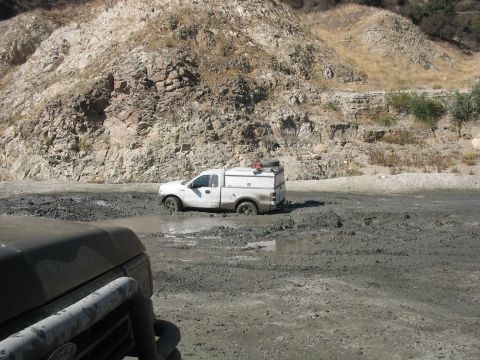 1987 Ford Bronco - White Ford F-150 stuck in mud, viewed from another vehicle in a rocky area.