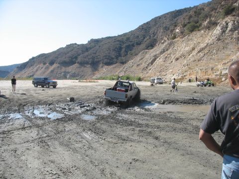 1987 Ford Bronco - Several off-road vehicles navigate a muddy terrain with a steep, rocky hillside in the background.