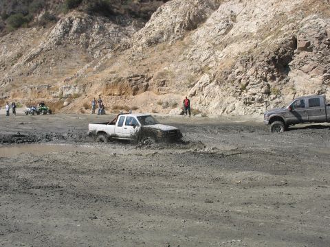 1987 Ford Bronco - Two pickup trucks navigate a muddy terrain with steep, rocky hills in the background.