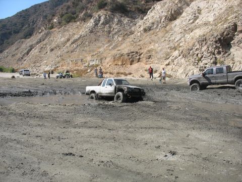 1987 Ford Bronco - Several vehicles are stuck in a muddy area with hills in the background.