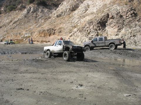 1987 Ford Bronco - Two pickup trucks navigate a muddy off-road course with steep, rocky hills in the background.