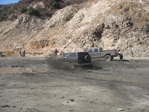 1987 Ford Bronco - A black 1987 Ford Bronco navigates a muddy off-road course, with a pickup truck and several onlookers present.