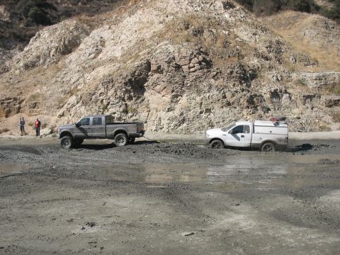 1987 Ford Bronco - Two trucks are stuck in a muddy area with a rocky hillside in the background.