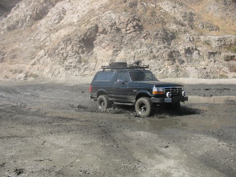 1987 Ford Bronco - Black Ford Bronco navigates a muddy track with a rocky hillside in the background.