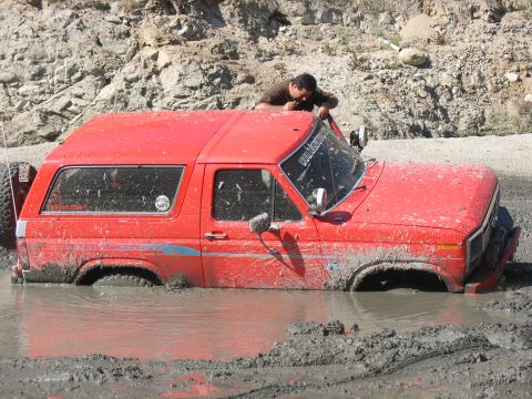 1987 Ford Bronco - A red SUV is deeply stuck in thick mud, with a person leaning over the roof.