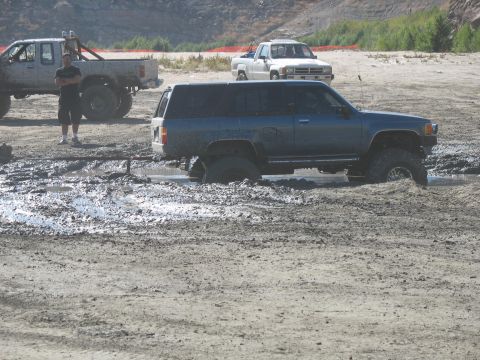 1987 Ford Bronco - A blue SUV navigates a muddy off-road course, with other trucks visible in the background.