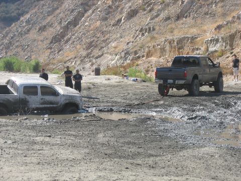 1987 Ford Bronco - Two trucks are stuck in a muddy, off-road environment with people observing.