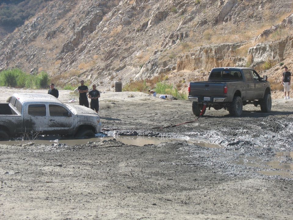 Mud-covered white truck submerged in mud, with a dark grey pickup truck nearby in a muddy, hilly landscape