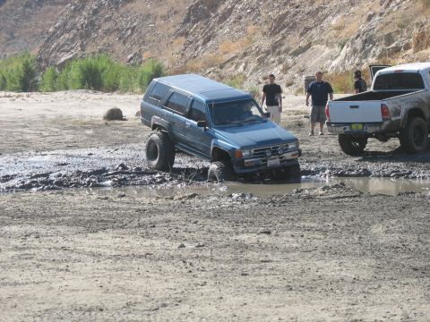 1987 Ford Bronco - A blue SUV navigates a muddy obstacle course, with another truck visible in the background.