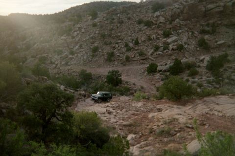 1979 Ford Bronco - A dark SUV navigates a rough, rocky terrain in a desert canyon under a hazy sky.