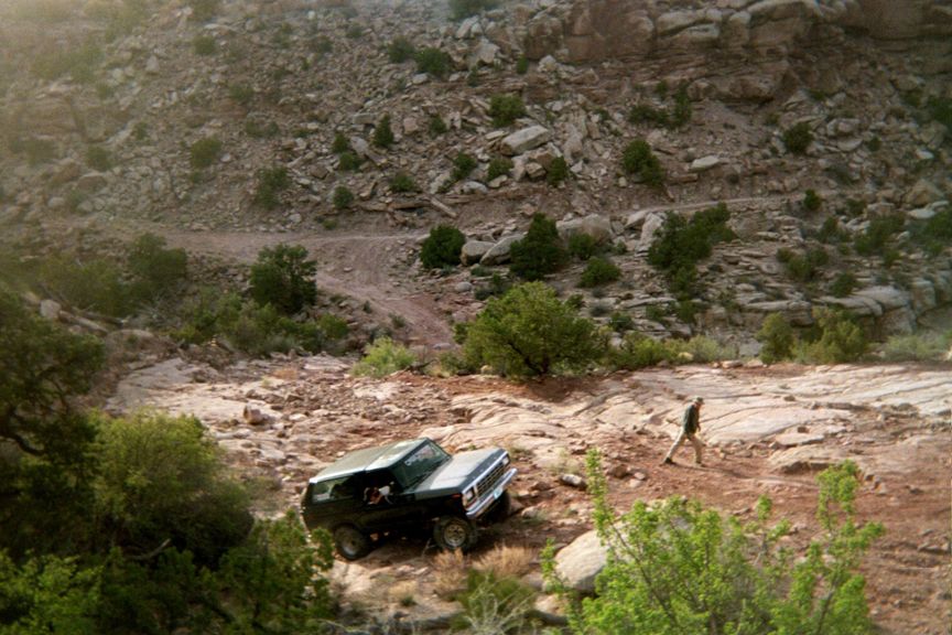 Dark SUV on rocky trail, person walking. Appears to be a 1978 Ford Bronco