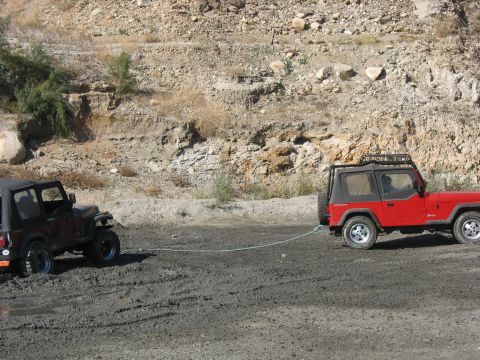 1987 Ford Bronco - A red Jeep tows a stuck black Jeep out of a muddy area, with a rocky hillside in the background.