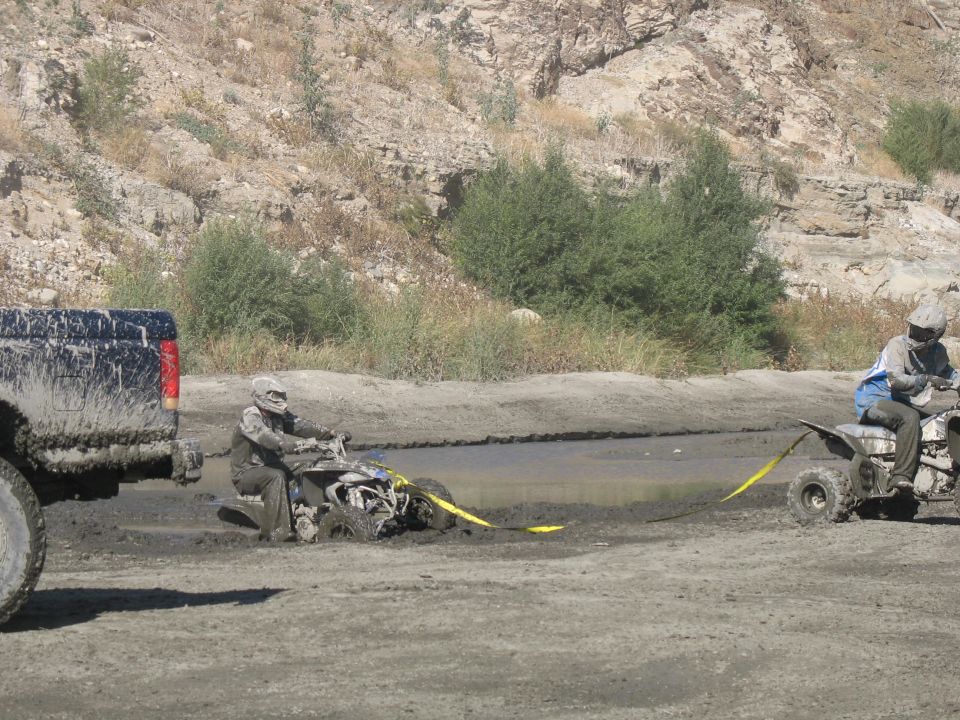 Muddy truck towing a stuck ATV out of a muddy ditch. Two people on ATVs are visible