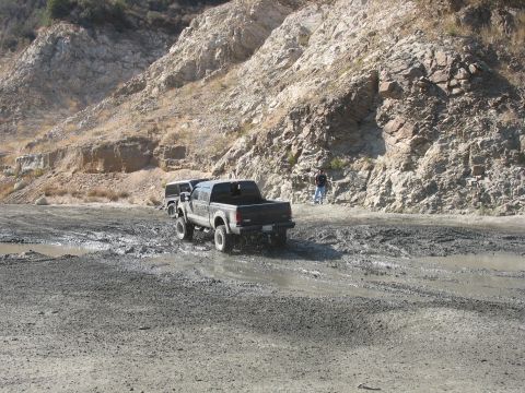 1987 Ford Bronco - Two trucks navigate a muddy terrain with a rocky hillside in the background.