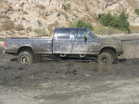 1987 Ford Bronco - Muddy crew cab pickup truck stuck in mud, side view. Appears to be a 1990s Ford F-Series.