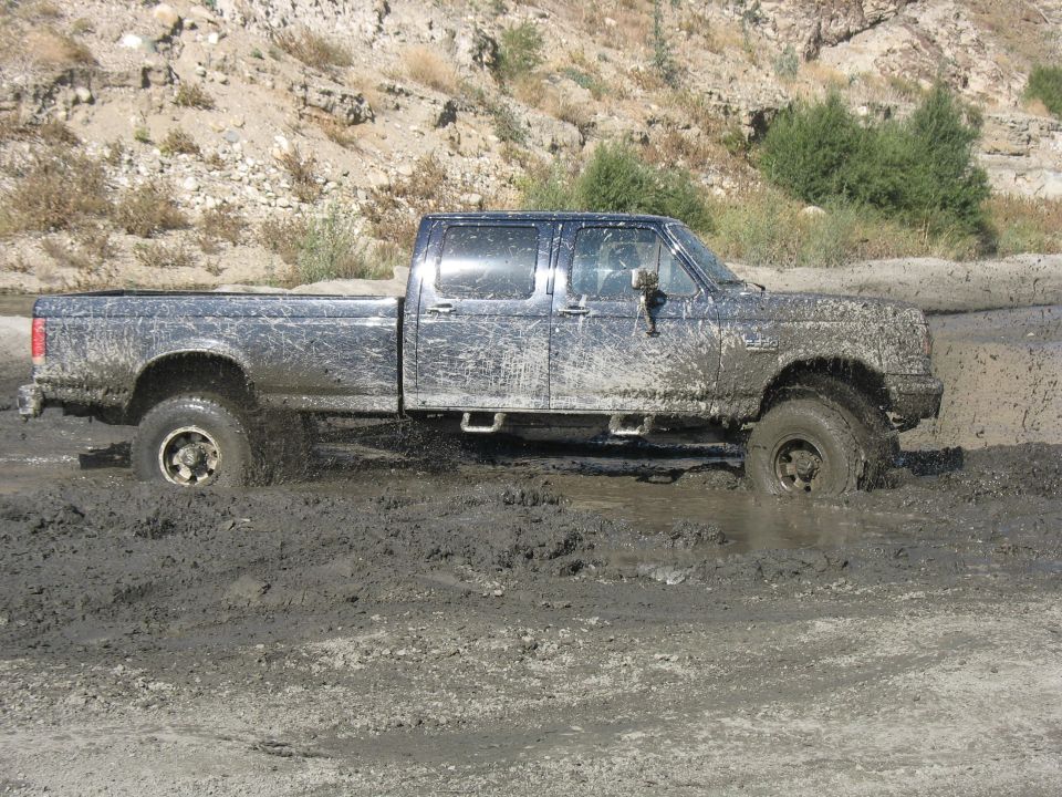Mud-covered crew cab pickup truck stuck in deep mud, side view. Appears to be a 1990s Ford F-Series