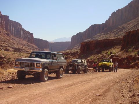 1979 Ford Bronco - Three off-road vehicles are parked on a dusty trail surrounded by canyon walls under a clear blue sky.