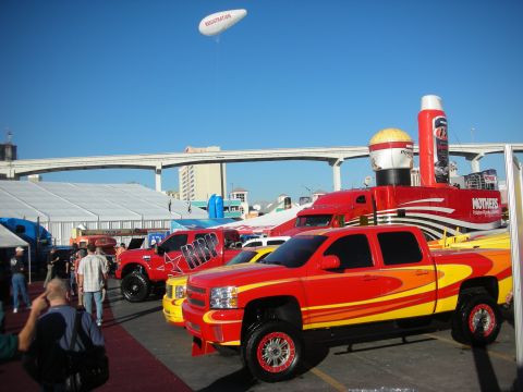 1998 Ford Crown Victoria - Customized lifted pickup trucks with flame and graphic paint parked under a clear sky at an outdoor automotive event near a freeway.