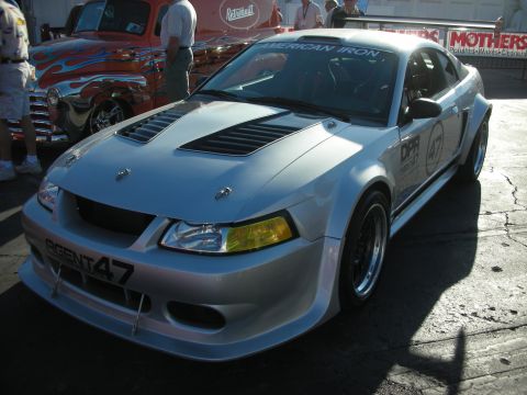 1998 Ford Crown Victoria - silver New Edge Ford Mustang race car with vented hood, wide fenders and American Iron and Agent 47 graphics at a track event.
