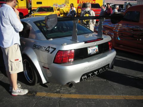 1998 Ford Crown Victoria - Rear of a silver Ford Mustang with a large adjustable wing, Agent 47 livery, and an Arizona plate at an outdoor show.