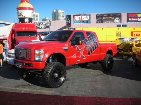 1998 Ford Crown Victoria - lifted red Ford Super Duty crew-cab pickup with black star graphics, oversized tires, and blue shocks on a show floor.