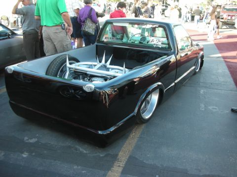 1998 Ford Crown Victoria - slammed black compact pickup with chrome wheels and a chromed engine mounted in the bed, parked among show crowds.