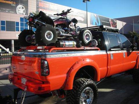 1998 Ford Crown Victoria - lifted orange Ford Super Duty pickup loaded with ATVs and dirt bikes in its bed, displayed outside a convention hall.