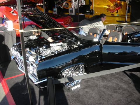 1998 Ford Crown Victoria - black convertible with its hood up reveals a polished engine on display behind ropes at an indoor automotive show.