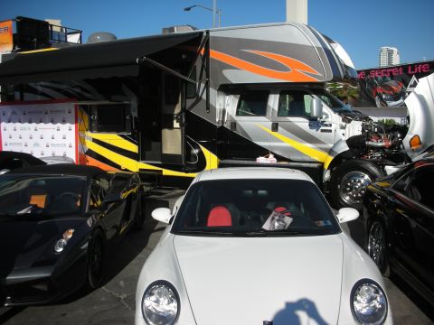 1998 Ford Crown Victoria - large graphic-wrapped RV motorhome towers behind a white sports car and a black sports car in an outdoor lot.