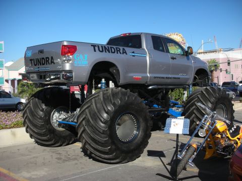 1998 Ford Crown Victoria - silver Toyota Tundra lifted onto enormous mud tires is displayed as a monster truck on a city street.