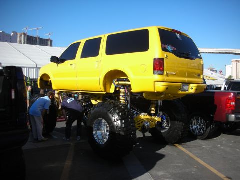 1998 Ford Crown Victoria - yellow Ford Excursion SUV raised on monster tires and a tall lift kit is displayed in an outdoor parking lot.