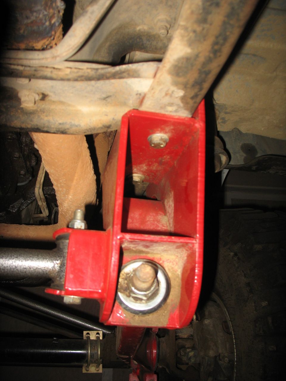 Red fabricated suspension bracket bolted to a rusty truck frame with large through-bolt