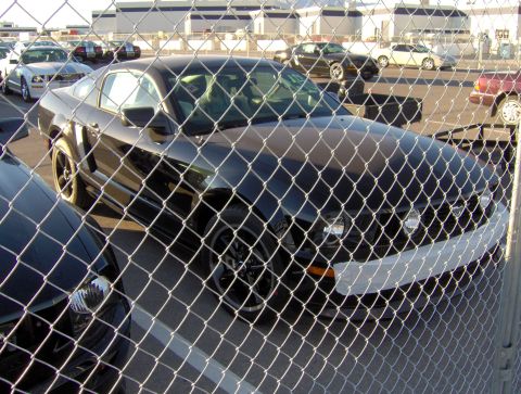 1978 Ford Bronco - A black Ford Mustang is parked behind a chain-link fence, partially obscuring its front end and a white bumper cover.