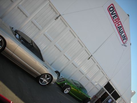 1998 Ford Crown Victoria - Angled shot of a silver car and a green car parked next to a white event trailer marked with the Overhaulin logo.