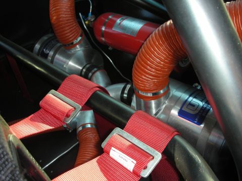 1998 Ford Crown Victoria - Close-up of a race car engine bay showing red harness webbing, orange cooling ducts, polished tubes, and a fire-suppression bottle.