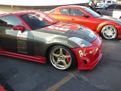 1998 Ford Crown Victoria - gray-and-red Nissan 350Z covered in sponsor decals and gold wheels sits beside an orange coupe at an outdoor show.