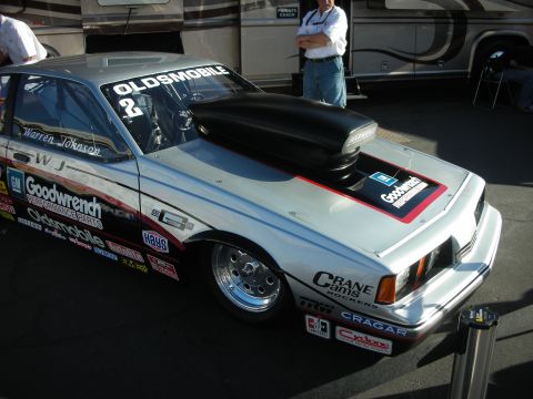 1998 Ford Crown Victoria - white Oldsmobile Pro Stock drag race car with a black hood scoop and Goodwrench and Crane Cams decals sits in a paddock.