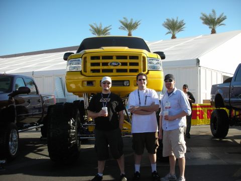 1998 Ford Crown Victoria - Three men stand for a photo in front of a yellow lifted Ford pickup raised high on oversized tires at an outdoor show.