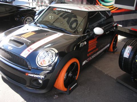 1998 Ford Crown Victoria - black MINI Cooper covered in Hankook tire livery sits on orange wheels beside a stack of tires at a show.