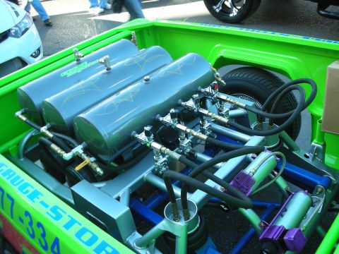 1998 Ford Crown Victoria - Chrome air-ride tanks, valves and lines fill the bed of a green show truck with a spare tire behind.